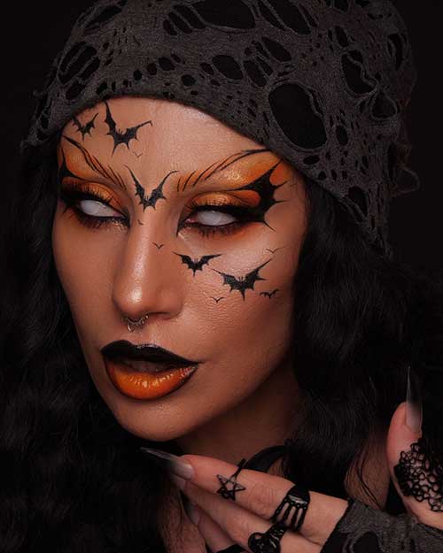 A Halloween makeup look showcasing burnt orange eyeshadow, black eyeliner, bats on the face, and contrasting lip colors.