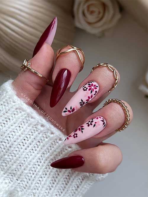 Elegant long burgundy almond nails with two nude pink accents, showcasing intricate floral and leaf designs in black and dusty rose.