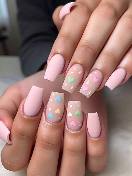 Long matte light pink coffin nails with 3D hearts in blue, pink, green, and yellow, adorned with white stars on a nude base.