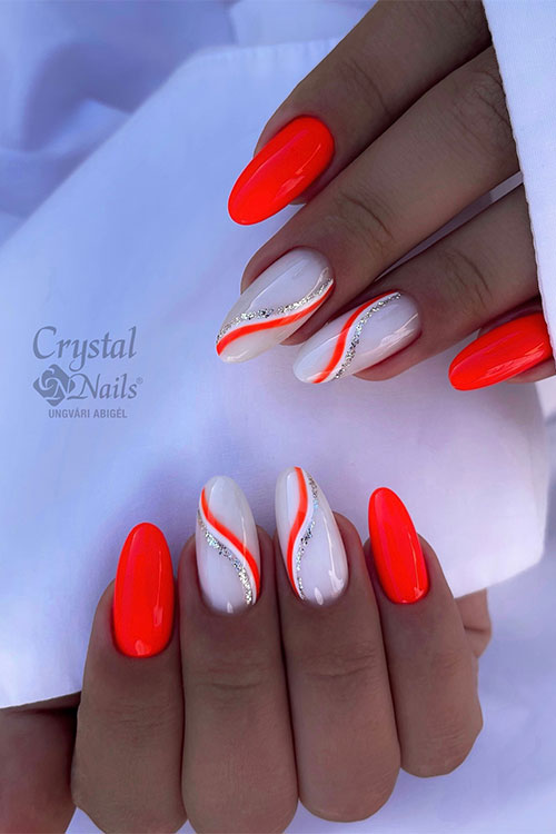 Long almond-shaped neon orange nails with two accent milky white nails adorned with white, silver glitter, and orange swirls