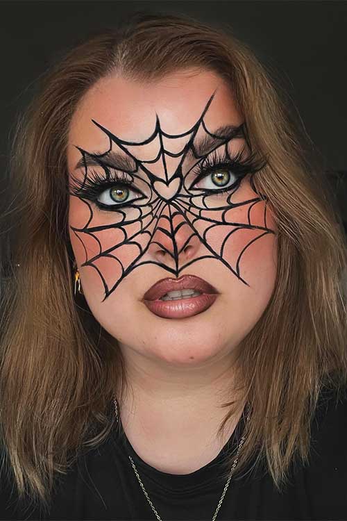 A large cobweb Halloween makeup envelops the face, highlighting warm brown lips beneath its intricate design.