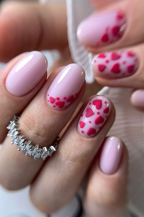 Short glossy nude pink nails with two accent nails adorned with red hearts and dots