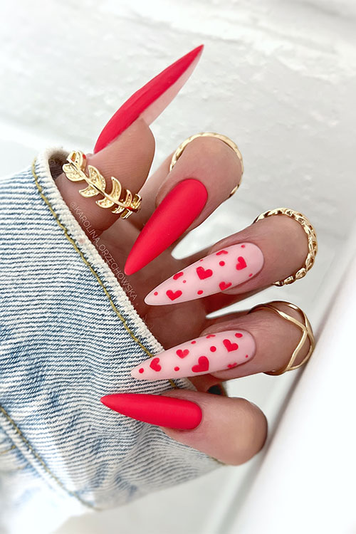 Stiletto matte red Valentine’s Day nails with two nude pink accent nails adorned with tiny red heart shapes