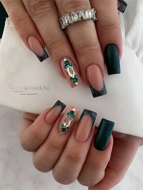 Long square dark green nails with glittery dark green French tips and a nude accent nail adorned with rhinestones.