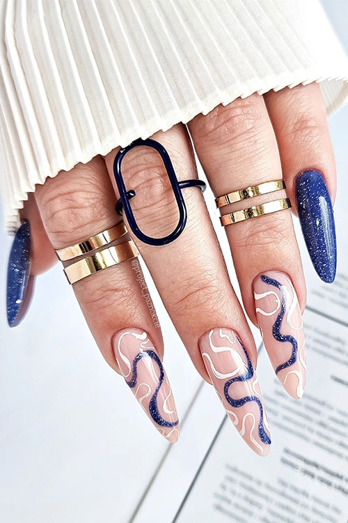 Long almond-shaped neon dark blue nails with glitter with three accent nude nails adorned with glitter blue and white swirls