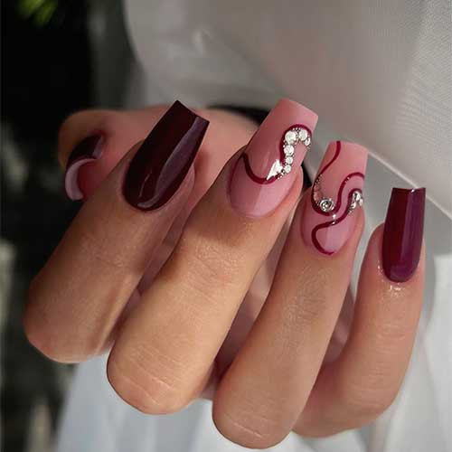 Burgundy coffin nails with two nude pink accent nails featuring elegant burgundy swirls and sparkling crystals.