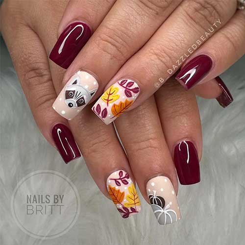 Fall burgundy nails featuring autumn designs like maple leaves, pumpkins, and a raccoon face on accent nails.