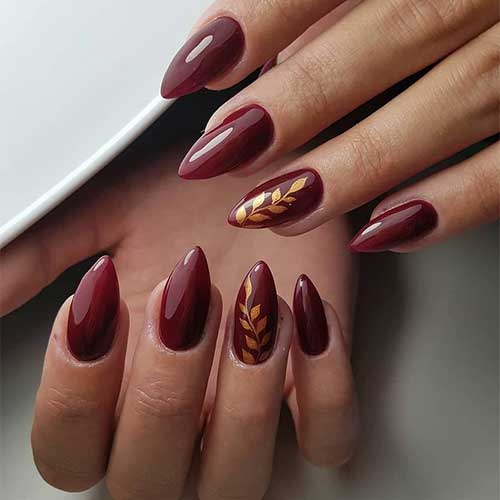 Glossy almond burgundy nails featuring a gold leaf design on an accent nail, showcasing elegance and creativity.