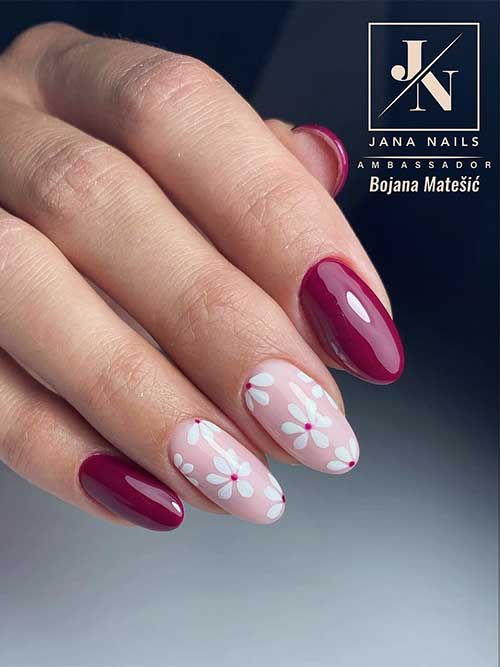 Shiny burgundy nails embellished with white flowers, alongside two accent nails painted in a subtle nude pink color.