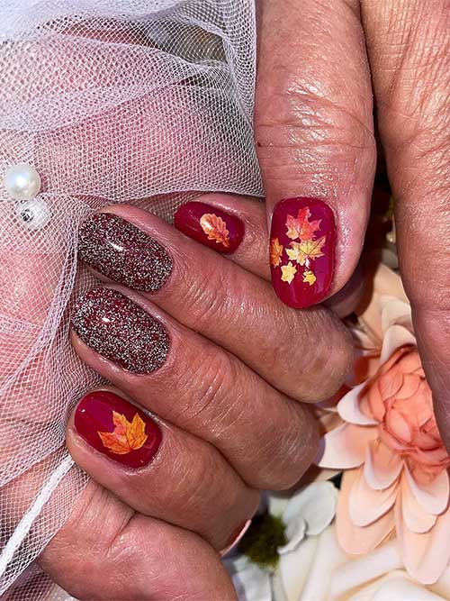 Short burgundy nails adorned with maple leaf stickers and two accent nails featuring glittery burgundy polish.