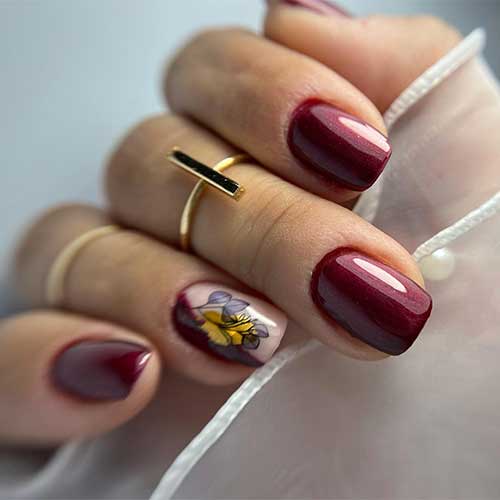 Short burgundy gel nails featuring fall leaves on a negative space accent nail, perfect for autumn-inspired nail art.