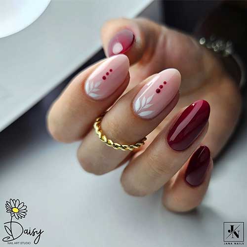 Simple burgundy nails with two nude pink accents featuring white leaf designs and burgundy dots for a stylish look.