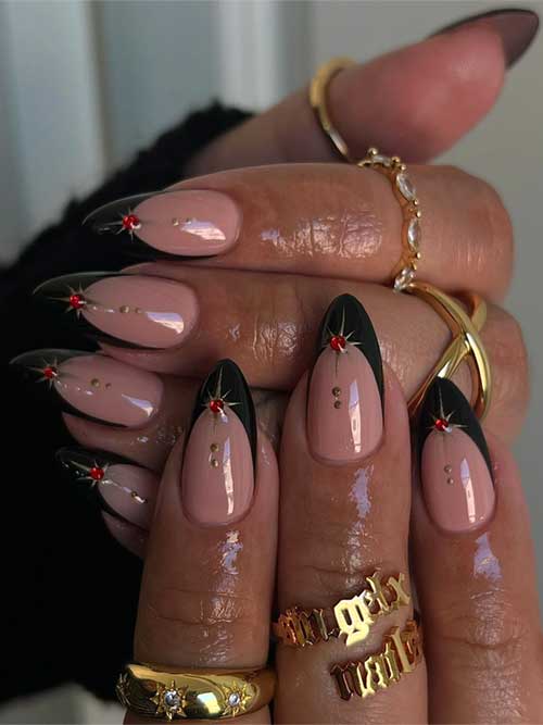 Black French tip nails on a nude base, adorned with gold stars and red rhinestones, featuring gold dots on the nude area.