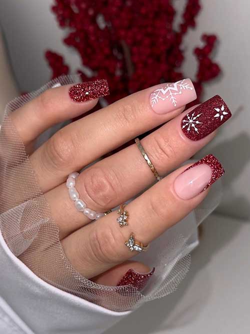 Dark red glitter winter nails 2024 decorated with snowflakes and featuring a French accent and another nude accent nail decorated with a big white snowflake.