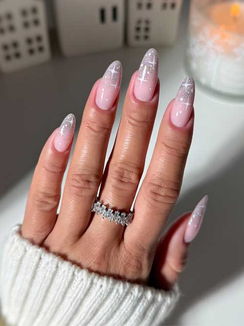 Long almond-shaped silver glitter ombre nails on a nude pink base, decorated with dots and snowflakes for a festive look.