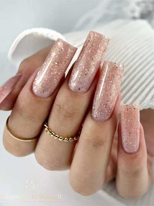 Long nude nails adorned with gold glitter and small black shreds for an elegant and stylish appearance.