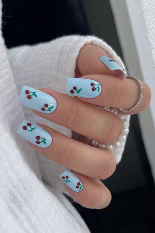 Light pastel blue nails with delicate red cherry designs and green leaves, embodying the beauty of spring blooms.