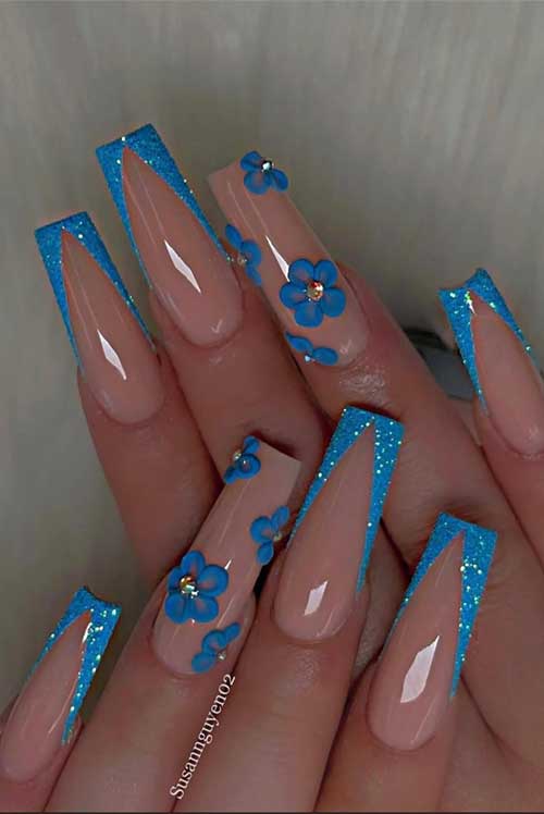 Nude coffin nails with striking blue glitter tips in a V-shape and charming 3D blue flowers with rhinestones for a spring touch.