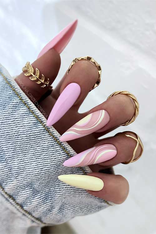 Long, pointed nails in pink, yellow, and swirls, adorned with gold rings, paired with a denim jacket for a chic look.