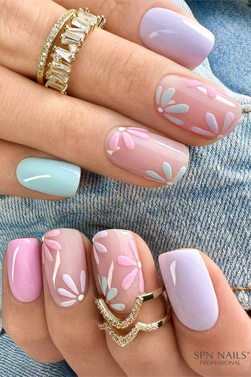 Pastel spring nails featuring soft blue, lavender, pink, and nude hues with floral patterns and gold rings for a chic look.